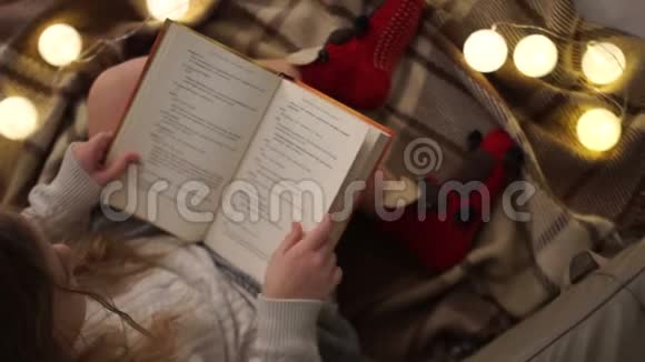 圣诞节的晚上女孩坐在铺着毯子的床上看书看圣诞灯看家庭视频的预览图