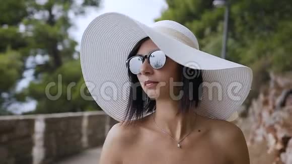 一个戴着白色帽子和太阳镜的漂亮黑发女人在海边的公园里散步戴帽子的漂亮女孩的肖像视频的预览图