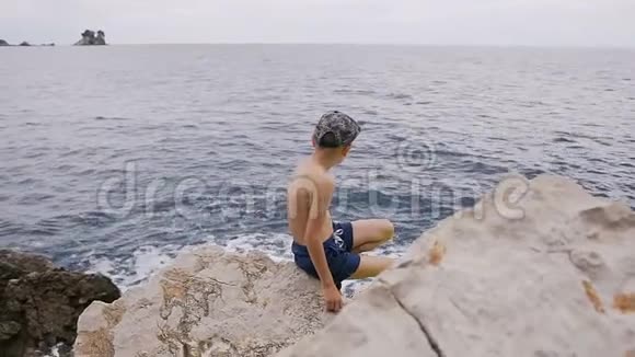 后景阳光明媚的一天穿着带帽子的短裤的小男孩坐在岩石湾的石滩上小孩子看着视频的预览图