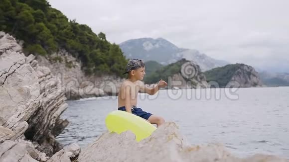 七八岁的男孩坐在岩石上看着一个美丽的地平线可爱的男孩带着充气戒指坐在上面视频的预览图