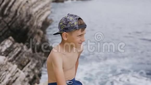 夏天或度假时戴着帽子的帅哥坐在海和岩石背景上的岩石上可爱的男孩视频的预览图