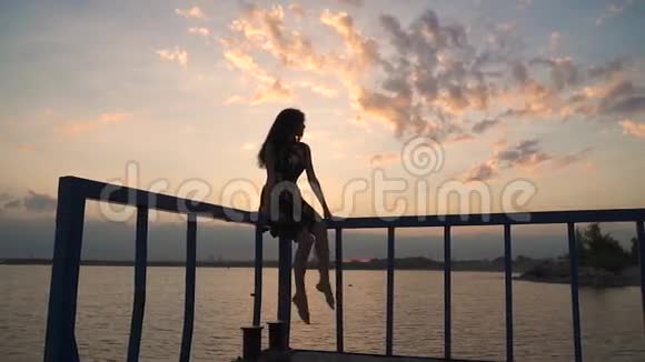 日落时分美丽的女孩坐在码头上摆动着一条的腿一个穿着浅色衣服的漂亮女孩的剪影视频的预览图
