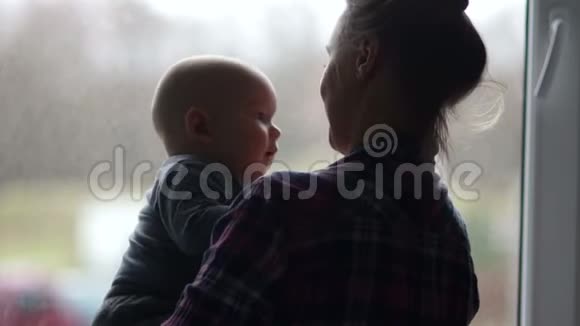一位年轻母亲带着孩子的剪影一个年轻的女人小心翼翼地抱着她的孩子看着窗外等待视频的预览图