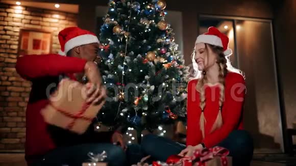 幸福的一对夫妇正坐在家里客厅的一块蓬松的地毯上一个男人正在给女孩送圣诞礼物视频的预览图