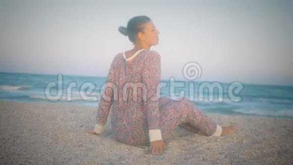 穿着睡衣的女孩伸展身体在日落前坐在海滩上视频的预览图