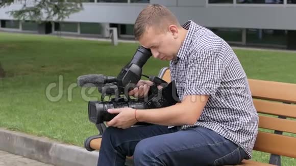 坐在大楼附近的长凳上的摄影师正在用电视摄像机拍摄视频的预览图