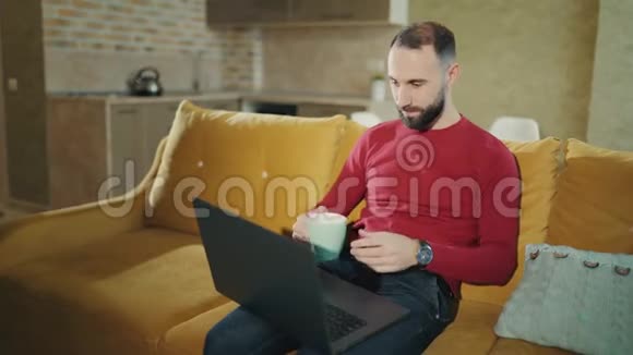 留胡子的人在家里用笔记本电脑时喝咖啡或茶会暂停4k个镜头视频的预览图