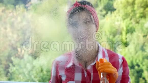 透过窗户观看微笑美丽的女人戴着手套通过喷洒清洁产品清洁窗户使用视频的预览图