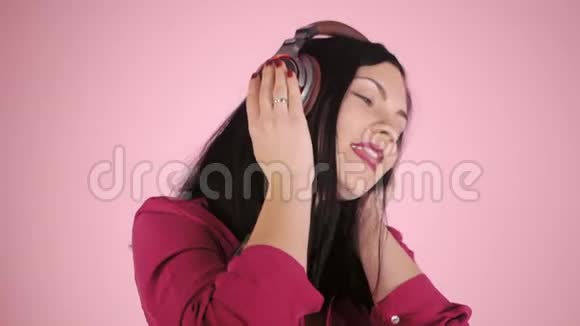 戴着耳机听着音乐的美女闭着眼睛站在衬衫上的粉色背景上视频的预览图
