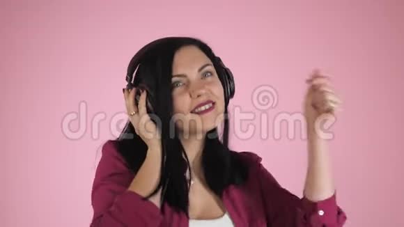 美丽的年轻女子戴着耳机听着音乐唱歌闭着眼睛站在衬衫上的粉红色背景上视频的预览图