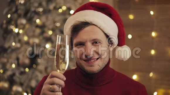 一个戴着圣诞帽拿着一杯香槟的帅哥看着圣诞树背景上的摄像机他举起了视频的预览图