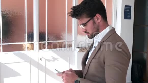 英俊的年轻商人一边坐在办公室的桌子上一边使用他的智能手机视频的预览图