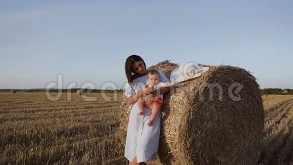 日落时带着儿子在田里的年轻女子一个穿着裙子的黑发可爱的女孩站在靠近稻草的田野里视频的预览图