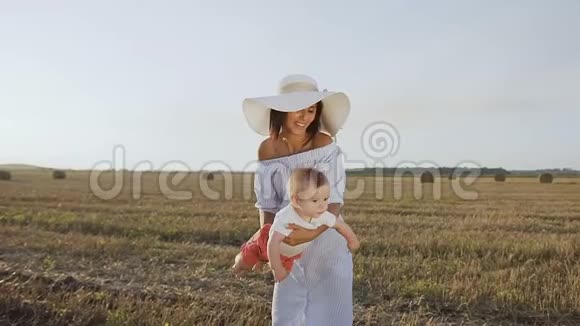 迷人的年轻女子穿着连衣裙和帽子正在与她的孩子在田野里玩日落妈妈和儿子一起玩视频的预览图