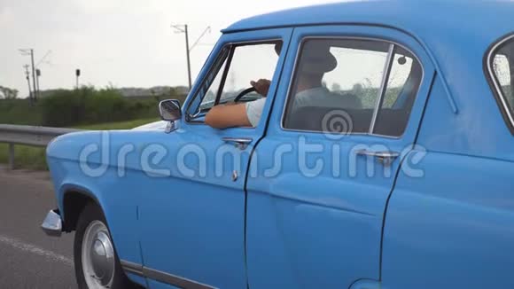 侧面看不可辨认的人戴着帽子驾驶蓝色复古车后雨在夏季旅行一个年轻人视频的预览图