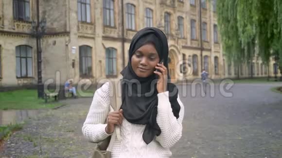 年轻的非洲穆斯林学生戴着头巾一边打电话一边微笑站在大学附近的公园里视频的预览图