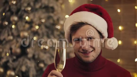 迷人的男人戴着圣诞帽的香槟看着圣诞树背景上的相机他举起了视频的预览图