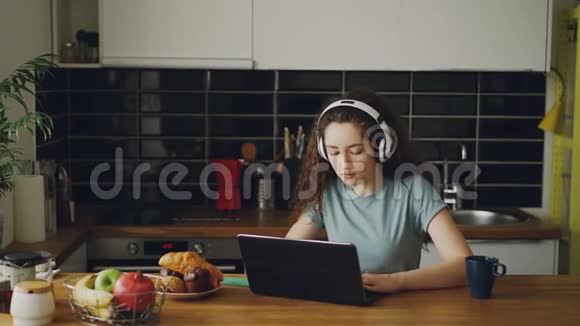 积极快乐的白种人漂亮的卷发少女坐在桌旁和笔记本电脑上听音乐的人聊天视频的预览图