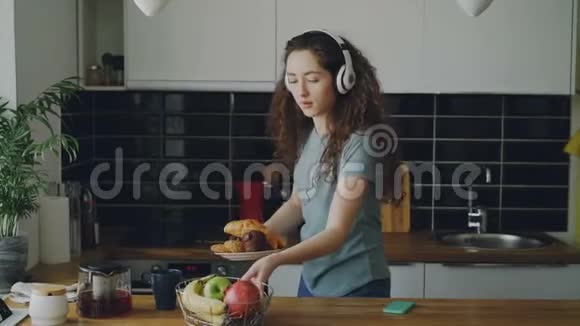 可爱的白种人少女戴耳机跳舞准备早餐喝咖啡放红色茶壶视频的预览图