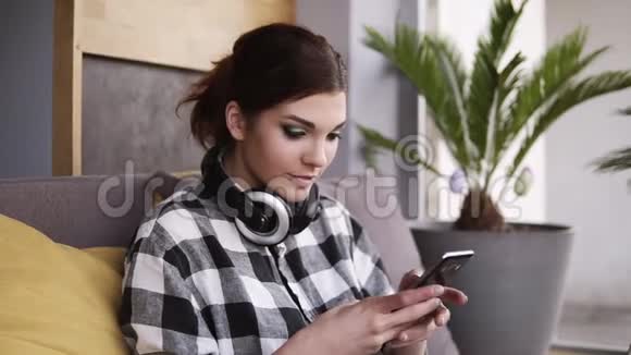 可爱的黑发女人坐在有黄色枕头的沙发上手里拿着手机滚动着微笑印度支视频的预览图