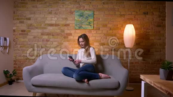 一位年轻迷人的白种人深色头发的女性在手机上浏览的特写照片她坐在室内的沙发上视频的预览图