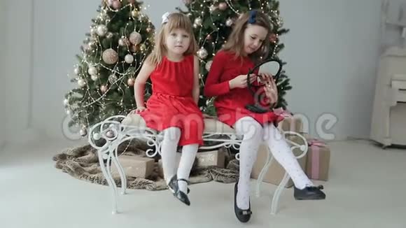 两个穿红色衣服的女孩坐在圣诞树附近的长凳上玩镜子圣诞节视频的预览图