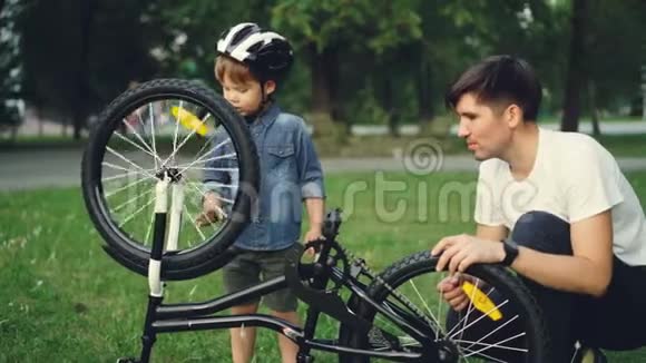 好奇的孩子戴着头盔一边旋转自行车轮和脚踏车一边父亲在公园的草坪上和他说话视频的预览图
