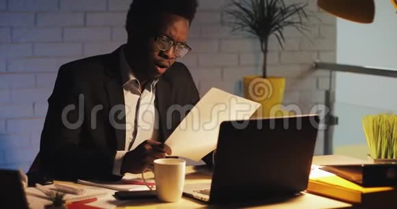 厌倦了年轻的黑人商人手提电脑和文件在夜间办公室工作他在处理文件视频的预览图