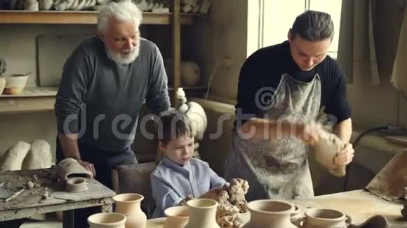 专业的陶工在家里的工作室里的工作台上捏黏土而他的儿子正在帮助他和年迈的父亲视频的预览图