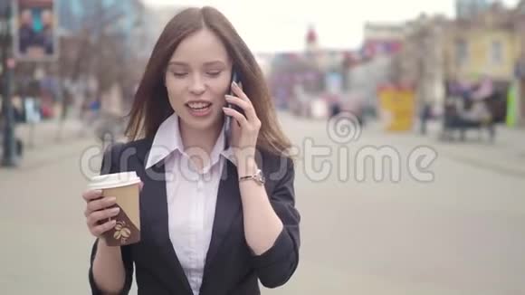 一幅严肃的女人穿着外套在电话里说话拿着咖啡的肖像背景上模糊的街道视频的预览图