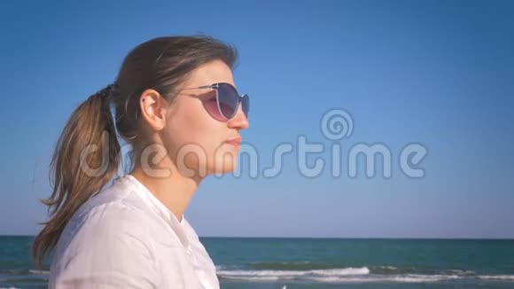 穿着白色衬衫和太阳镜的女孩从玻璃杯里啜着水微笑着坐在海边视频的预览图