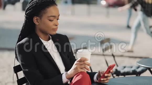 时尚的非洲商务女性使用智能手机喝咖啡坐在舒适的咖啡馆的夏季露台上通信视频的预览图