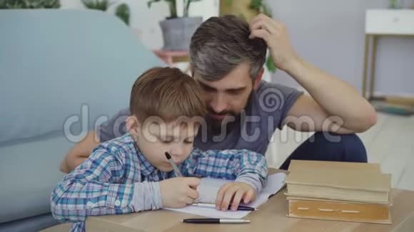 可爱的孩子在笔记本上写字他的父亲坐在桌子附近看着他的儿子学前教育视频的预览图