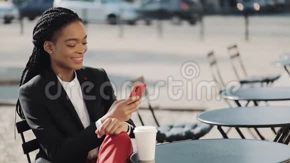 时尚的非洲商务女性使用智能手机喝咖啡坐在舒适的咖啡馆的夏季露台上通信视频的预览图