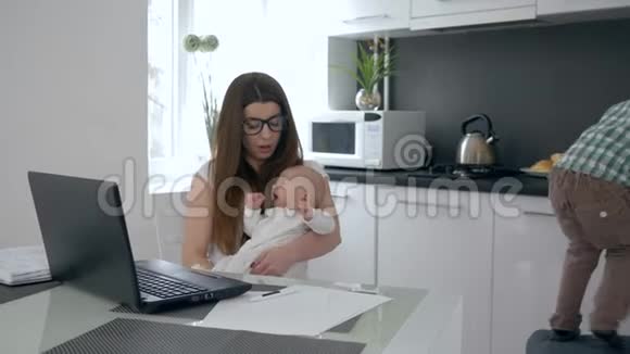 年轻的母亲坐在桌子上抱着哭泣的小男孩挨着她跳跃的大儿子厨房视频的预览图