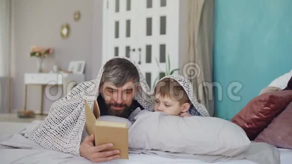 可爱的父亲和可爱的儿子正在读可怕的故事躲在毯子下躺在家里的床上快乐视频的预览图