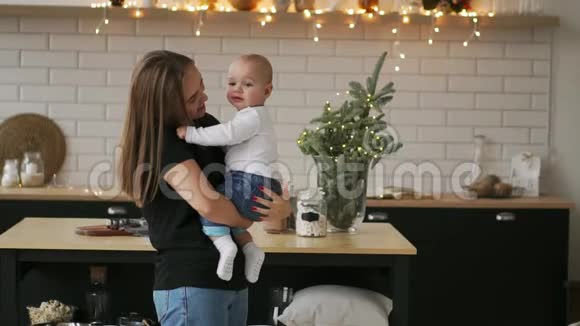 妈妈抱着宝宝孩子微笑着看着心爱的妈妈一起站在白色的厨房里过圣诞节视频的预览图