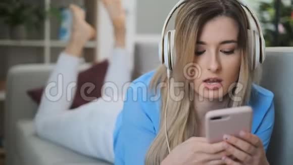 穿着蓝色服装在家欣赏音乐的年轻漂亮女孩女人从智能手机和耳机里听音乐视频的预览图