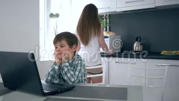 笔记本电脑后面的现代小孩在厨房的桌子旁看着一段等待早餐的视频视频的预览图