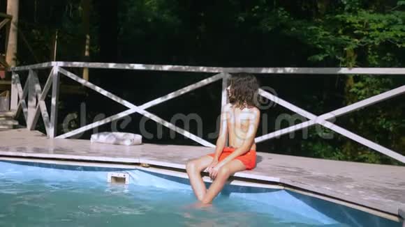 微笑的少年男孩卷曲的黑发坐在游泳池旁双腿在水里暑假假期视频的预览图