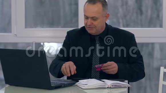老人把信用卡放在钱包里坐在桌子旁现代办公室里有笔记本电脑视频的预览图