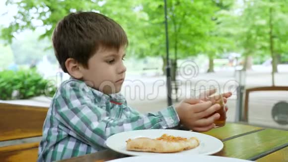不健康的食物小男孩从杯子里喝苹果汁在露天咖啡馆里吃一片披萨视频的预览图