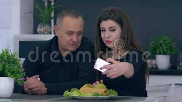 早餐女孩和爸爸坐在厨房的桌子上用手机自拍视频的预览图