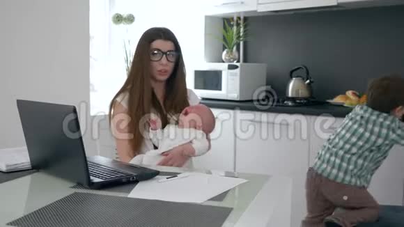 商业妈妈在笔记本电脑上工作并试图安抚厨房里哭泣的小儿子视频的预览图