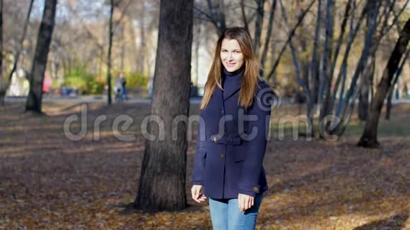 女人在树间快乐地跳跃秋天的叶子缓慢地移动4k60fps穿着外套的女孩在秋天的城市公园里散步视频的预览图