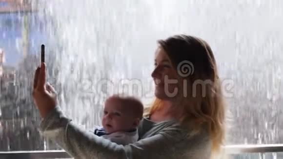 周末年轻妈妈抱着宝宝在水墙背景下用手机拍自拍照片视频的预览图