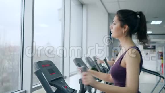 有氧运动训练年轻女性在有大窗户的健身中心用椭圆机做运动视频的预览图