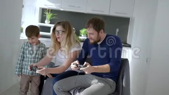 游戏玩家的家庭年轻的母亲和蹒跚学步的孩子和爸爸坐在房间的扶手椅上玩电子游戏视频的预览图