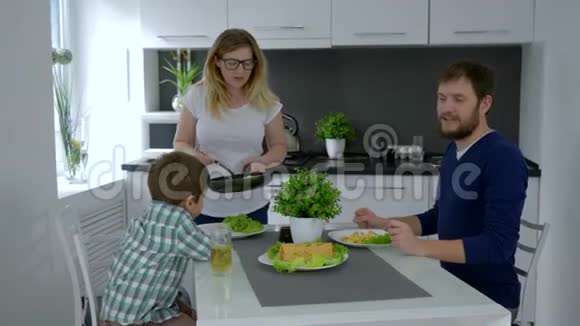 好妈妈用煎锅喂饭饿了的丈夫和儿子坐在餐桌上视频的预览图