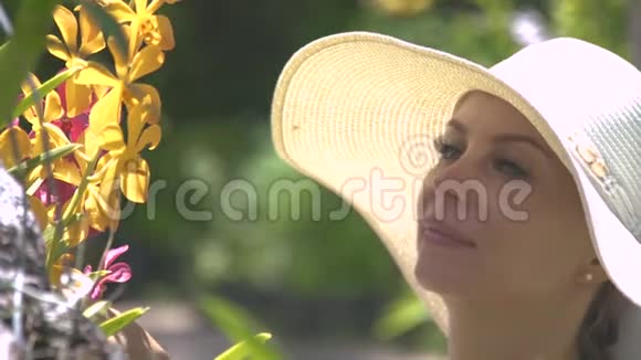 戴帽子的漂亮女人在夏天的花园里享受着盛开的兰花的香气女孩嗅着盛开的花朵的芬芳视频的预览图